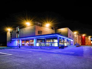 A modern hotel with a glass facade, a covered outdoor seating area, and a parking lot.