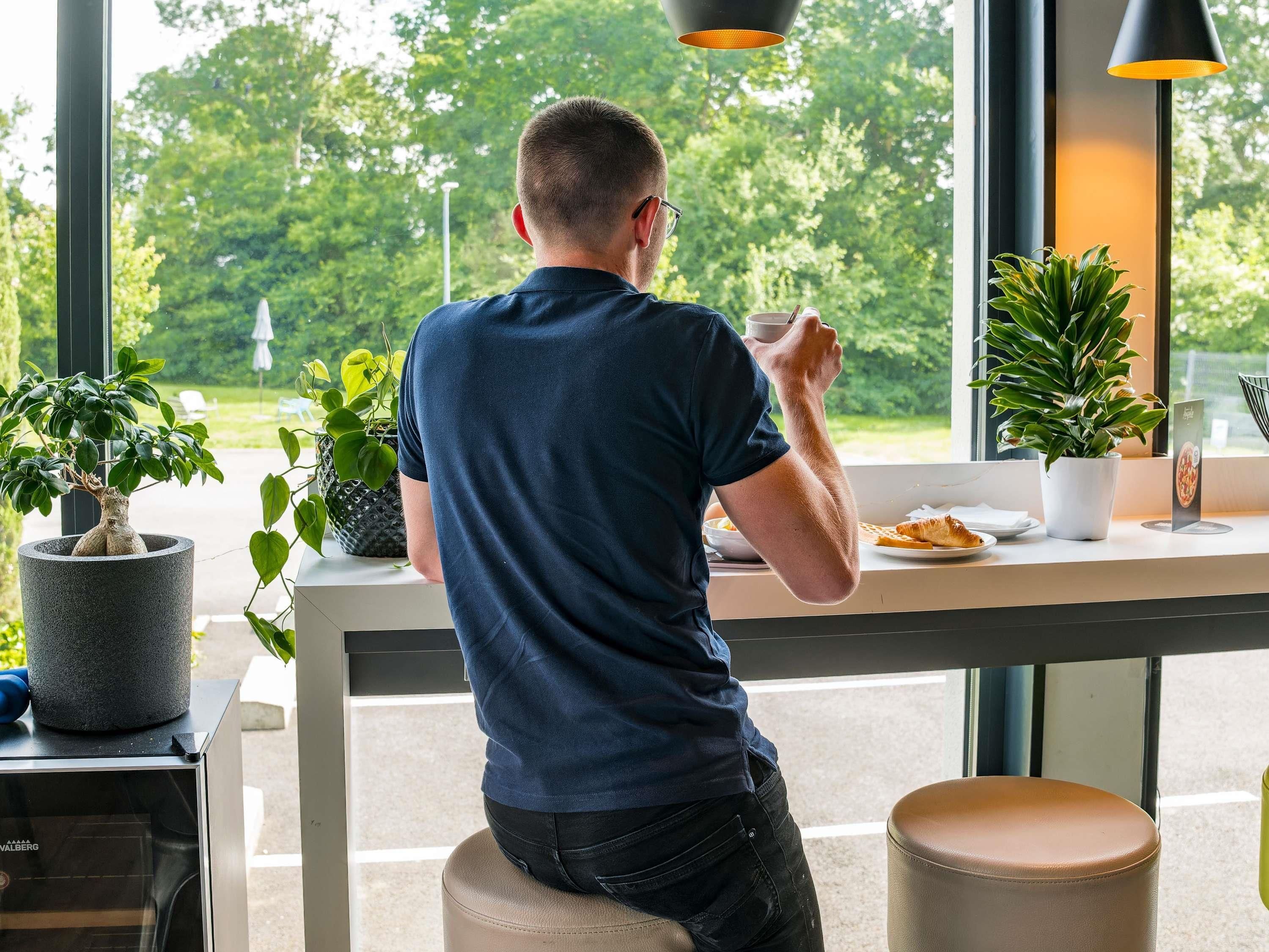 Una persona sentada a una mesa con vista a la vegetación exterior, una planta en maceta y un atril con menú.
