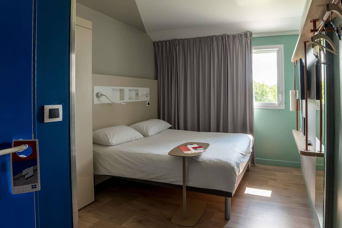 Ein Hotelzimmer mit einem Bett, einem Nachttisch, einem Fenster mit Vorhängen, einem wandmontierten Fernseher und einer Tür mit Griff.