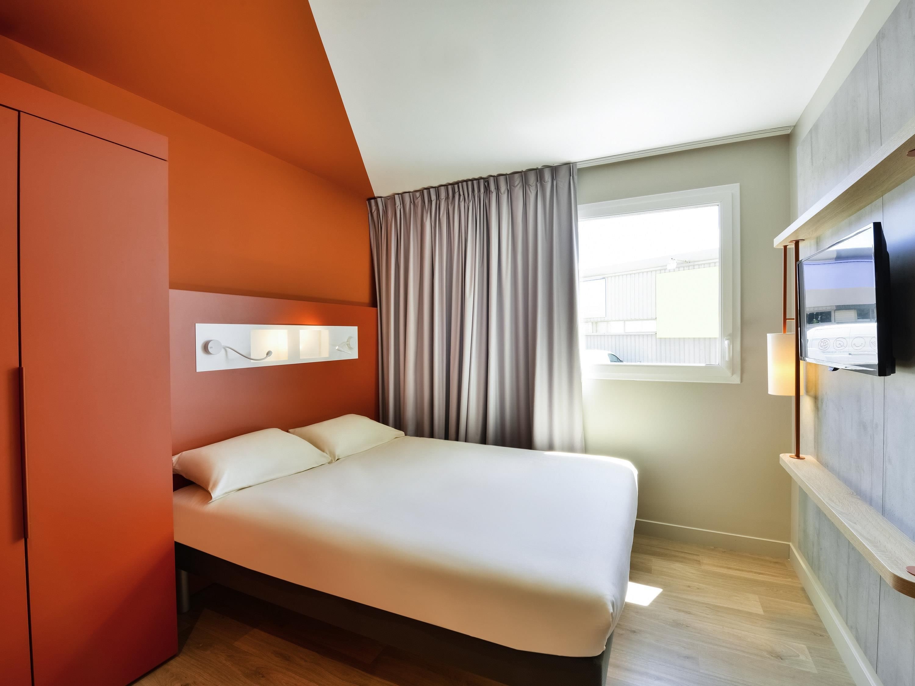 A modern hotel room with a bed, a TV mounted on the wall, a window with curtains, and a wardrobe.