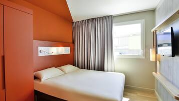 A modern hotel room with a bed, a TV mounted on the wall, a window with curtains, and a wardrobe.