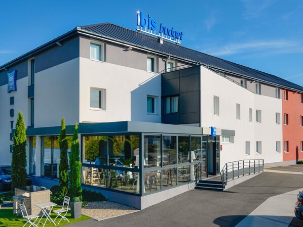 A modern hotel with a glass facade, outdoor seating area, and a sign reading "ibis budget". - ibis budget Mâcon Crêches (Chaintre)