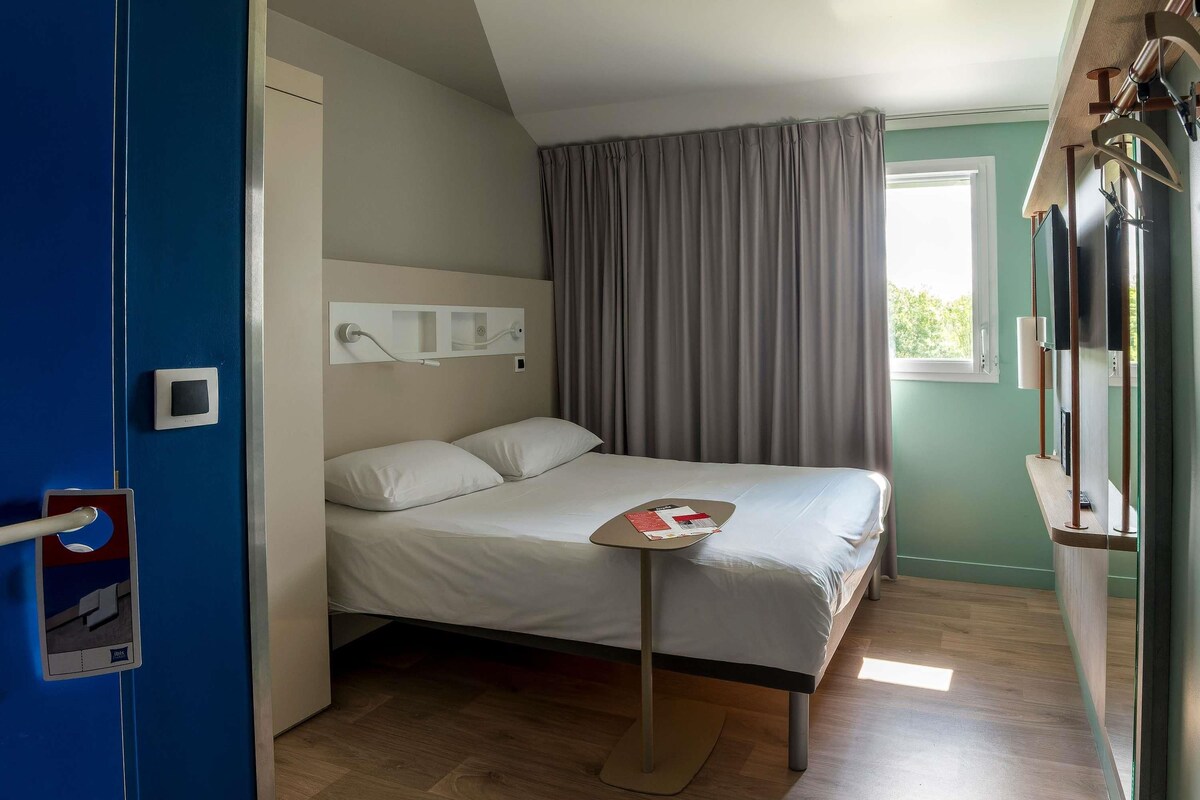 Una habitación de hotel con una cama, una mesita de noche, una ventana con cortinas, un televisor montado en la pared y una puerta con manija.