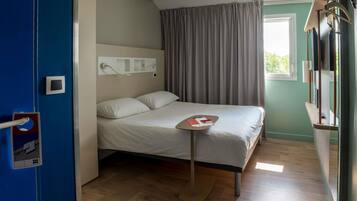 Een hotelkamer met een bed, nachtkastje, raam met gordijnen, een wandtelevisie en een deur met een deurklink.