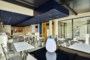 Un comedor moderno con mesas y sillas blancas, una lámpara esférica original y una cocina a la vista en el fondo.
