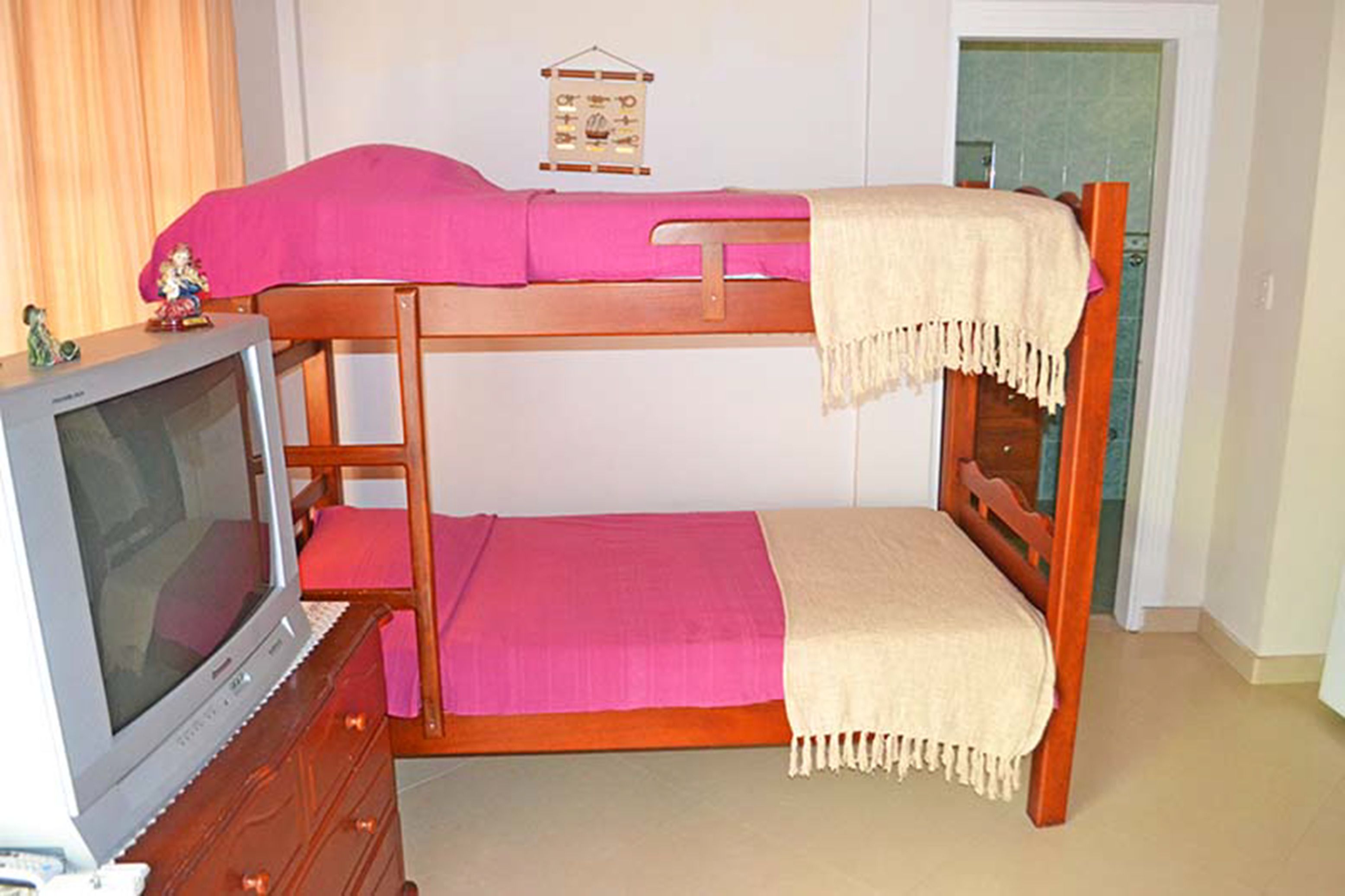 Un lit superposé avec une t élévision posée sur une commode, une couverture rose et un petit objet décoratif sur le lit du haut.