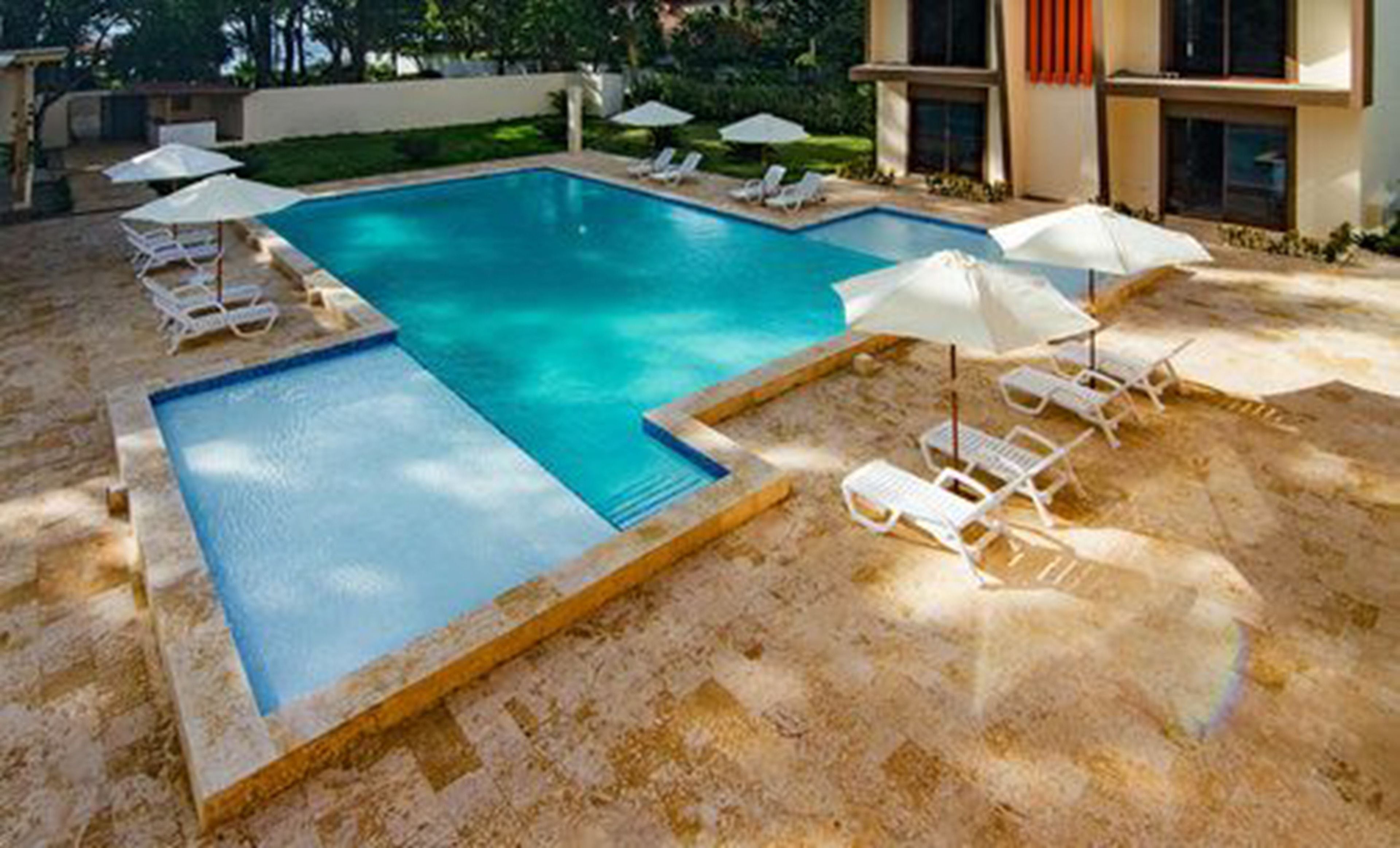 A rectangular swimming pool with a tiled deck, surrounded by white lounge chairs and umbrellas.