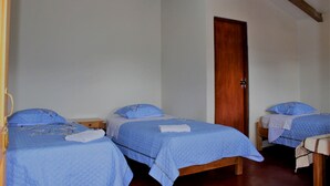 Une chambre avec trois lits simples, chacun recouvert d’une couverture bleue assortie à des coussins du même coloris. La chambre est dotée d’une porte en bois et d’un banc en bois.