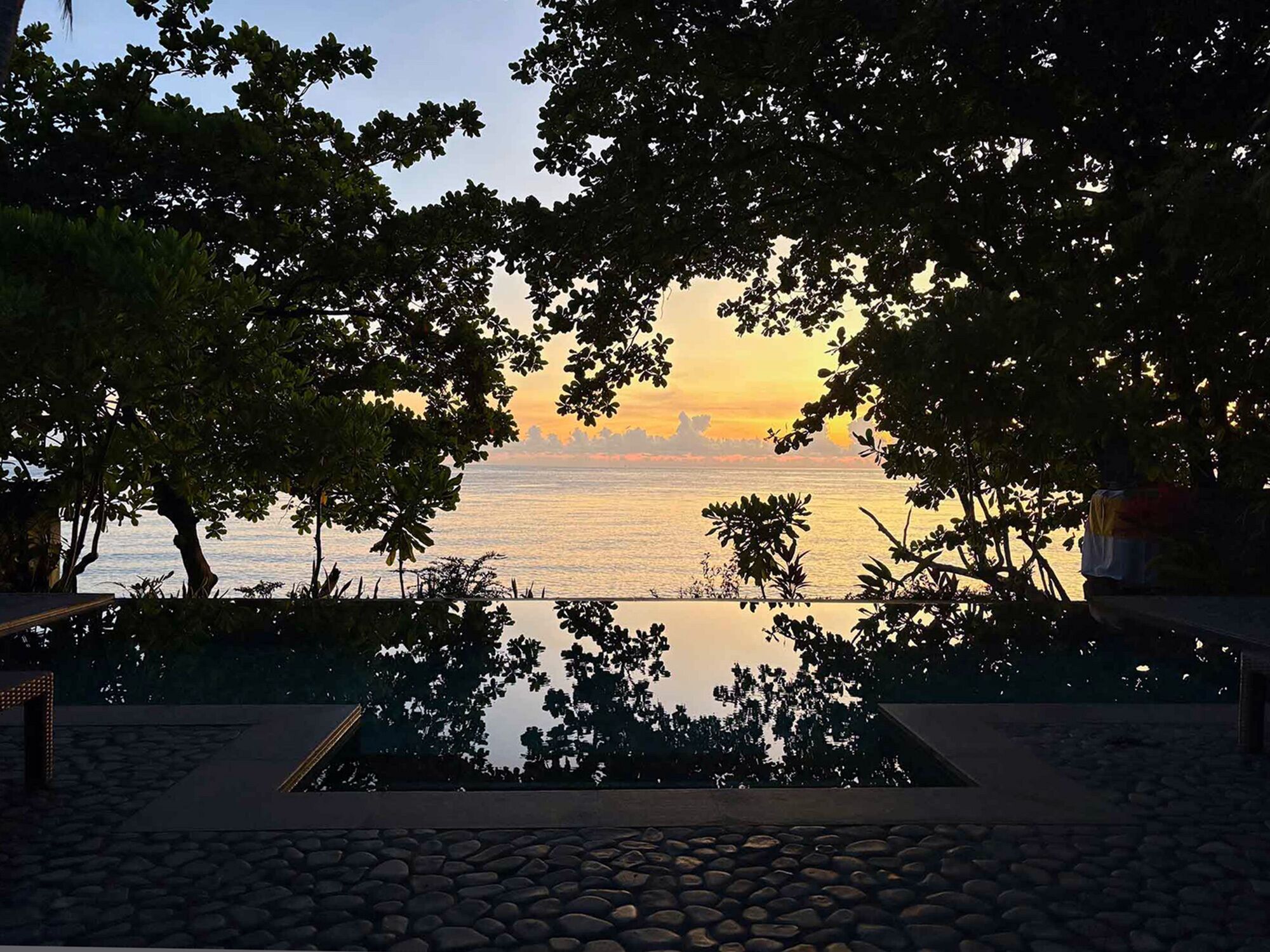 A serene outdoor pool area with a view of the sunset over the water, surrounded by trees.