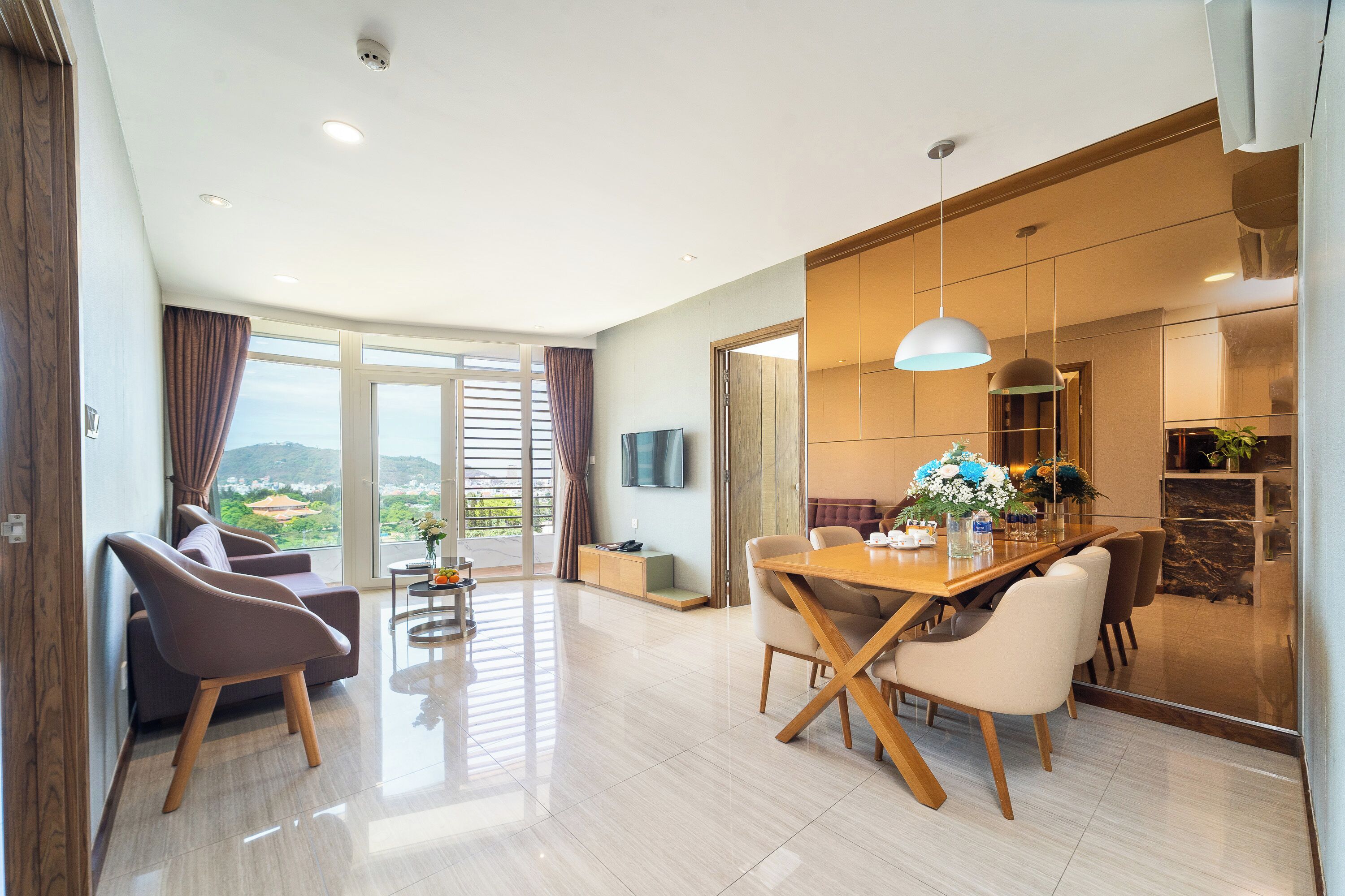Amplia sala de estar con una mesa de comedor, sillas y un sofá. Hay un televisor montado en la pared y un ventanal con cortinas.