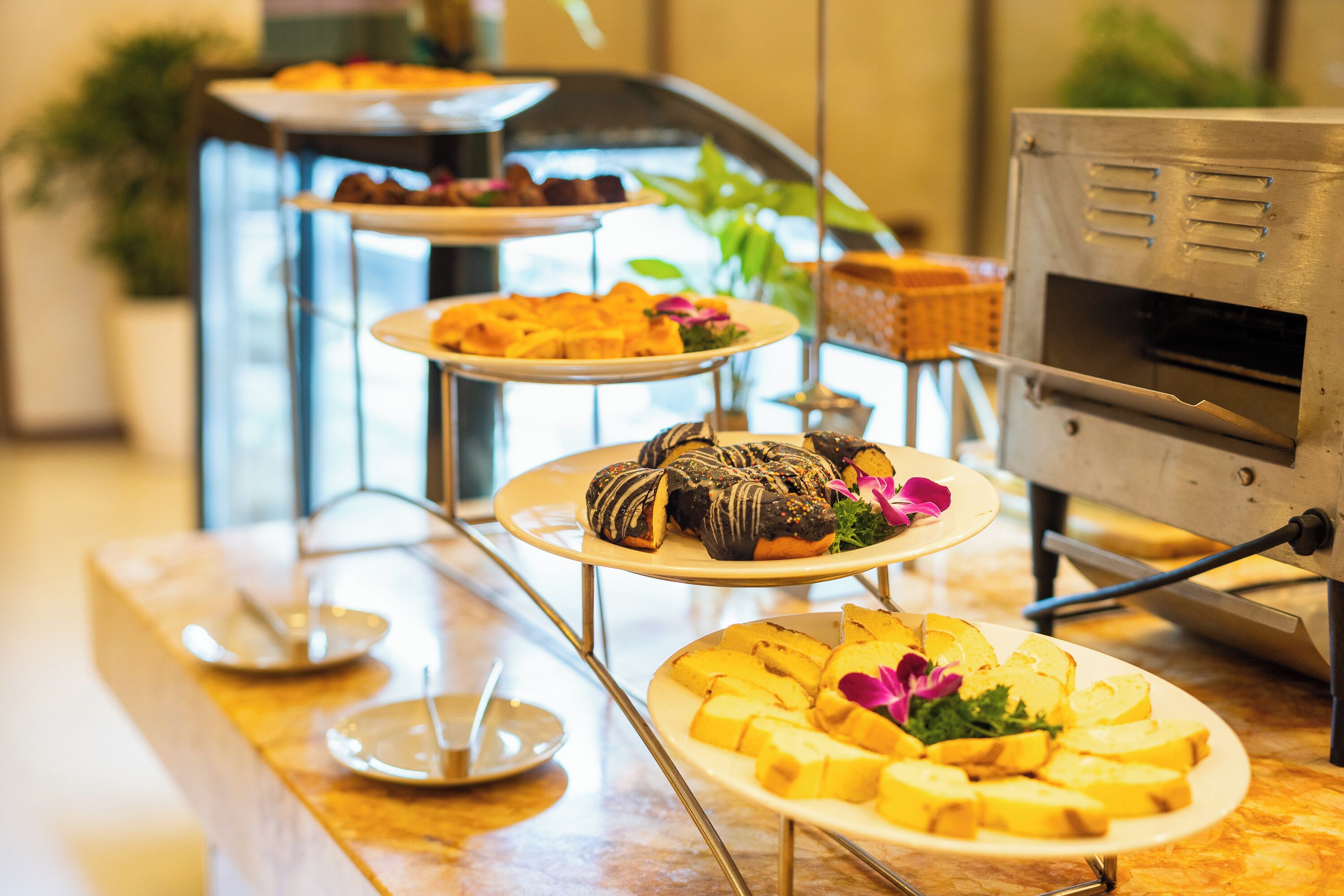 Un bufé con diversos alimentos en mesas escalonadas, incluyendo pasteles y pan, en un ambiente interior bien iluminado.
