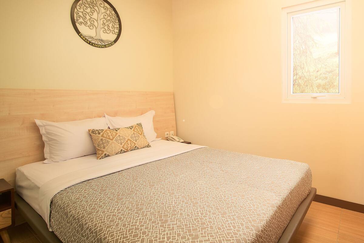 A neatly made bed with patterned bedding, a wooden headboard, a wall-mounted decorative item, and a window with a view.