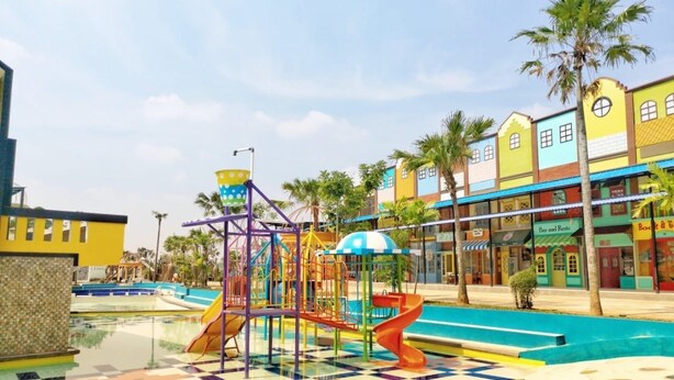 Un parque infantil con toboganes y estructuras para escalar, una zona de piscina con suelo de baldosas, y edificios coloridos con tiendas y restaurantes al fondo.