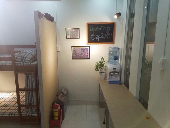A small room with a bunk bed, a desk with a water dispenser, a fire extinguisher, and a chalkboard sign indicating an opening date.