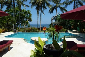 A pool area with lounge chairs and umbrellas, surrounded by palm trees and a view of the ocean. - Villa Damai (Seririt)