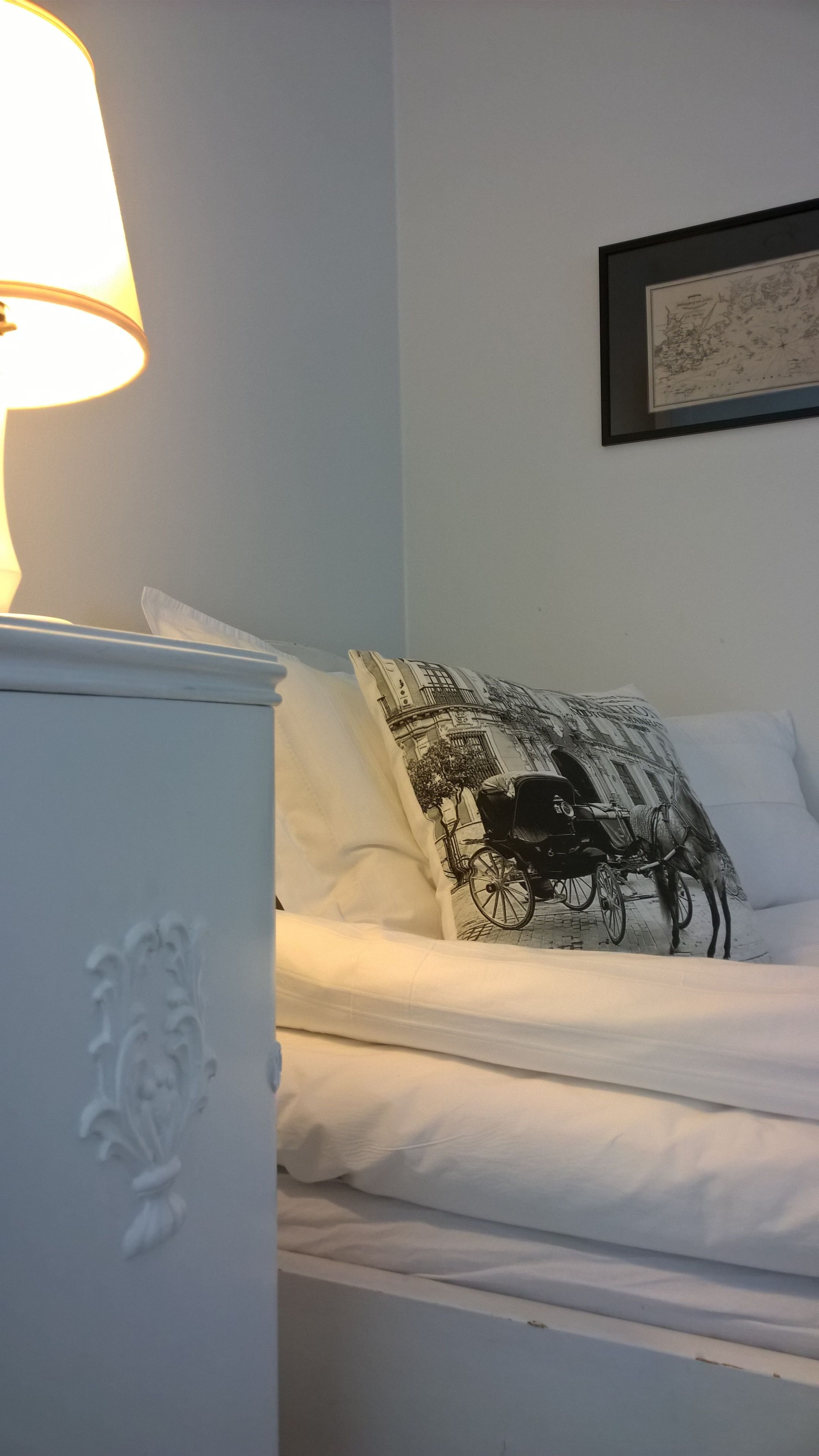Een net opgemaakt bed met witte lakens en een zwart-wit gestreept kussen. Links is een witte nachtkastje met een lamp te zien.