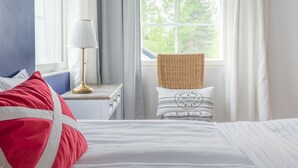 Un dormitorio con una cama, una mesita de noche con lámpara, una silla de mimbre y una ventana con cortinas.