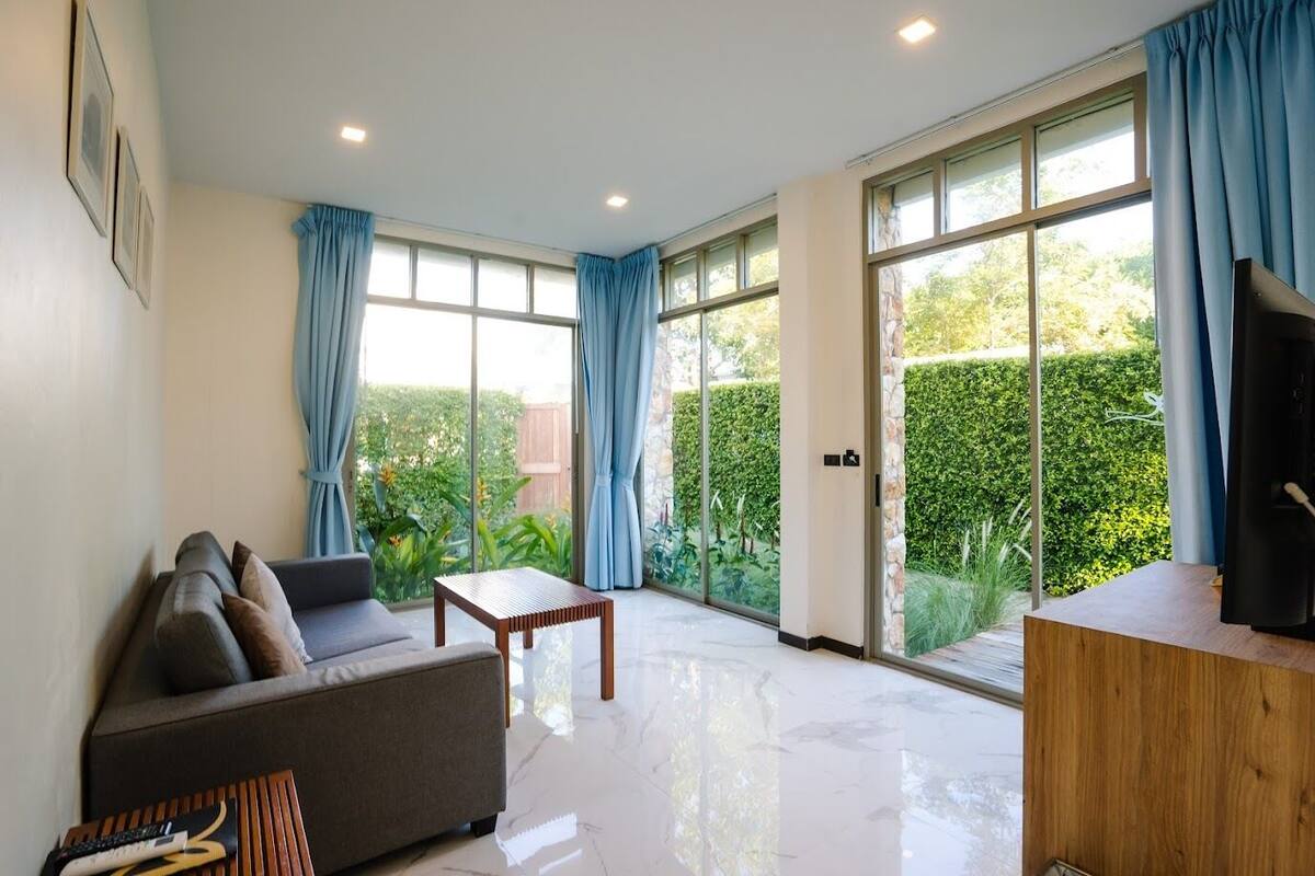 A modern living room with a sofa, a wooden coffee table, and a TV on a wooden stand. Large windows with blue curtains offer a view of a garden.