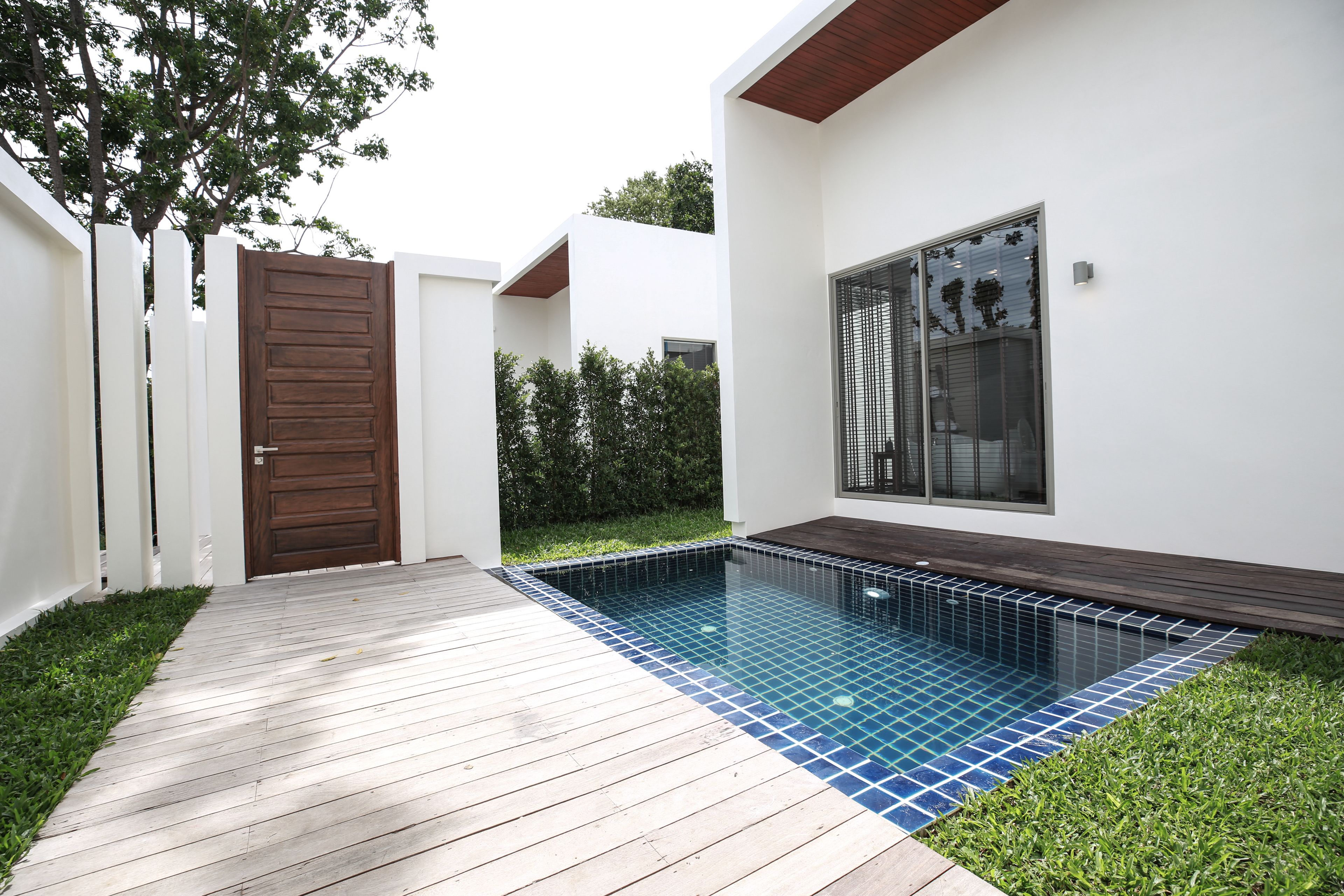 A modern outdoor swimming pool area with a wooden deck, a white building with a large window, and a brown gate.