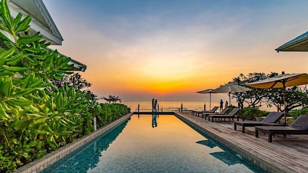 A poolside area with a view of the sunset over the ocean, lounge chairs, and umbrellas. - De Chaochom Hua Hin (Hua Hin)