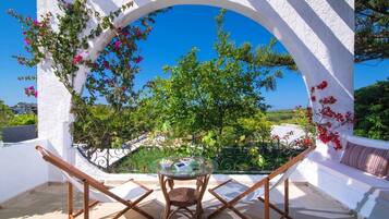 A white-walled outdoor seating area with a table and chairs, surrounded by greenery and a clear blue sky.