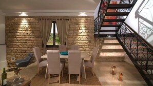 Uma sala de jantar com parede de pedra, uma mesa com cadeiras, uma escada com guarda-corpo de ferro forjado e duas velas acesas no chão.