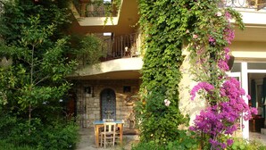 Un patio con un sendero de piedra, una mesa de madera y sillas. Se observa un balcón con barandilla de hierro forjado y un muro de piedra con una puerta.