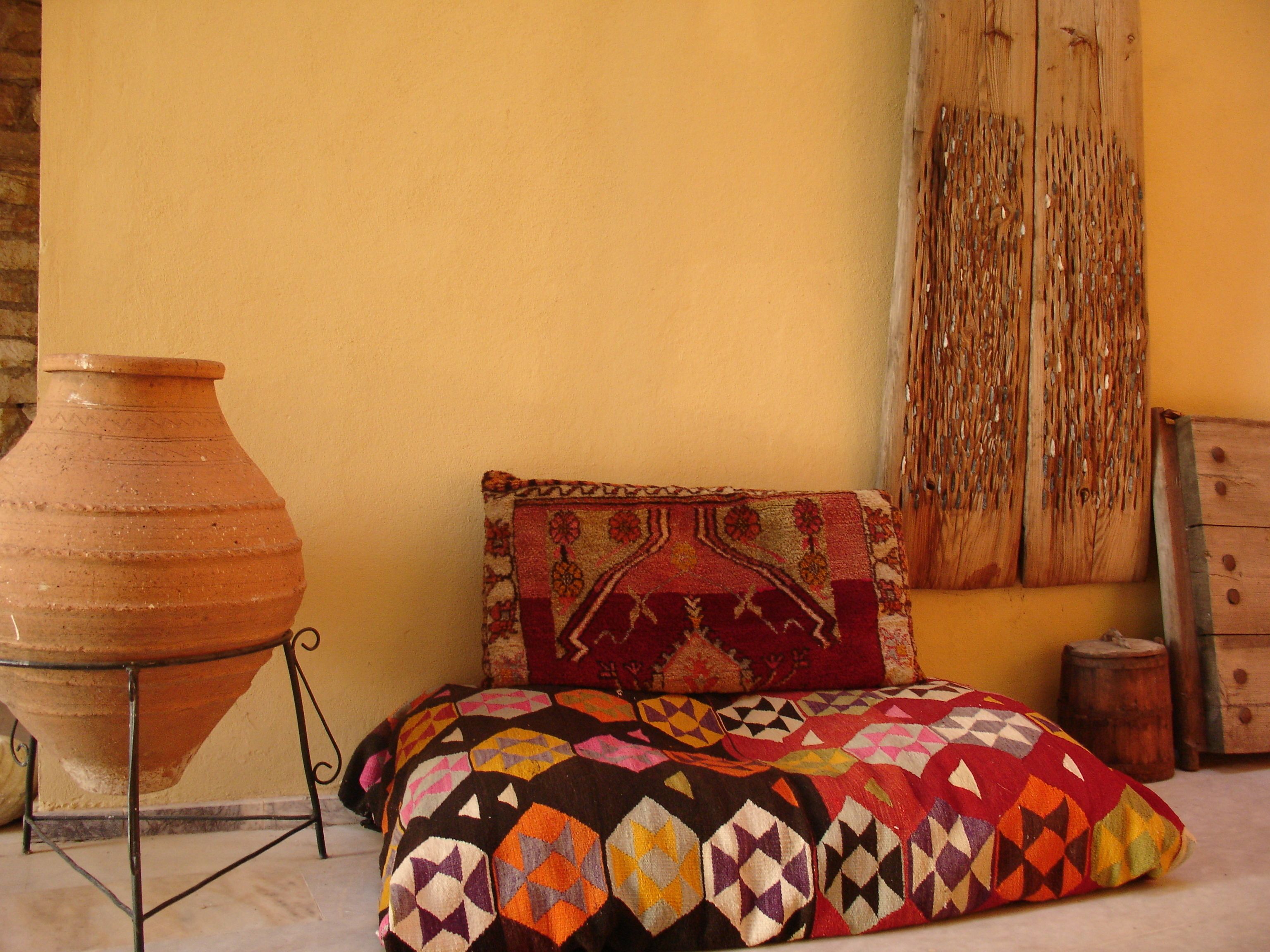 A room with a large clay pot on a stand, a colorful geometric-patterned cushion, and a wooden door with a woven curtain.