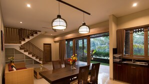 A spacious dining area with a large wooden table and chairs, a staircase with wooden railings, and large windows offering a view of greenery. - Niniku villa (Tegallalang)