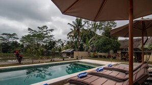 A poolside area with lounge chairs and umbrellas, surrounded by greenery and a building in the background. - Niniku villa (Tegallalang)