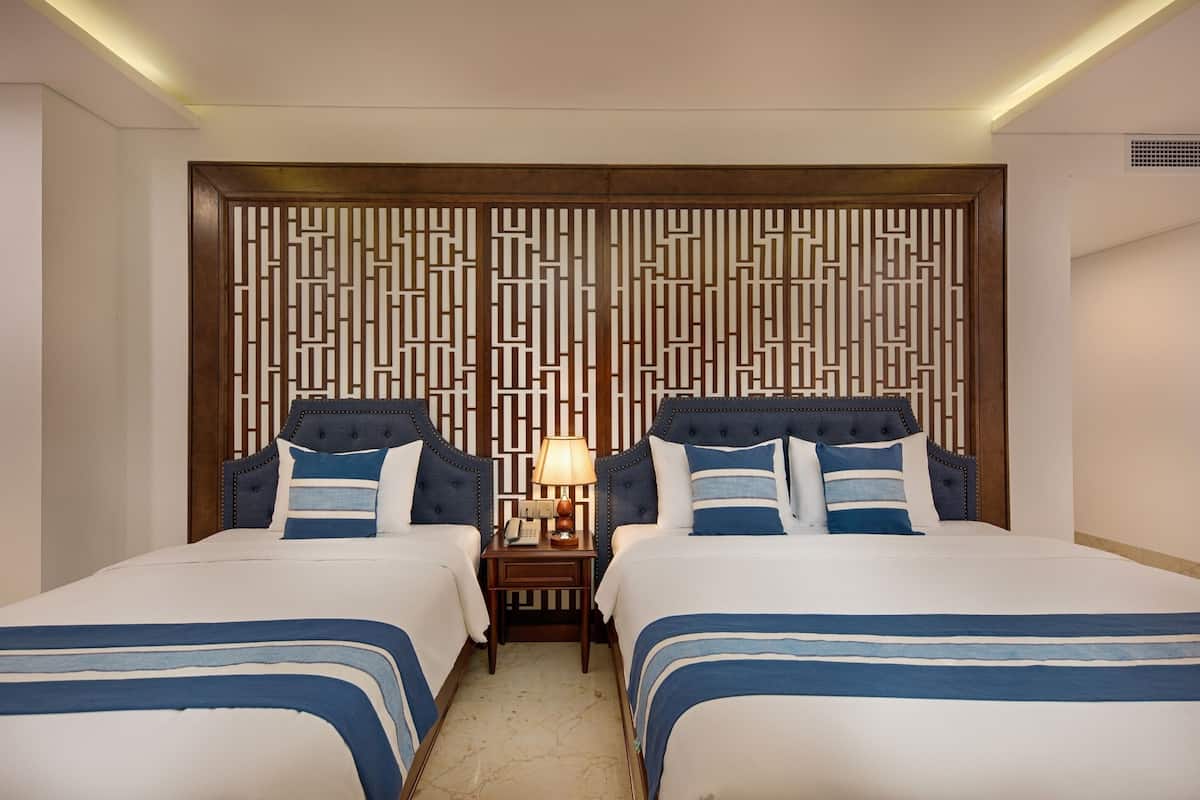 Une chambre d’hôtel avec deux lits, une tête de lit en bois ornée d’un motif géométrique, une table de chevet avec une lampe et un téléphone.