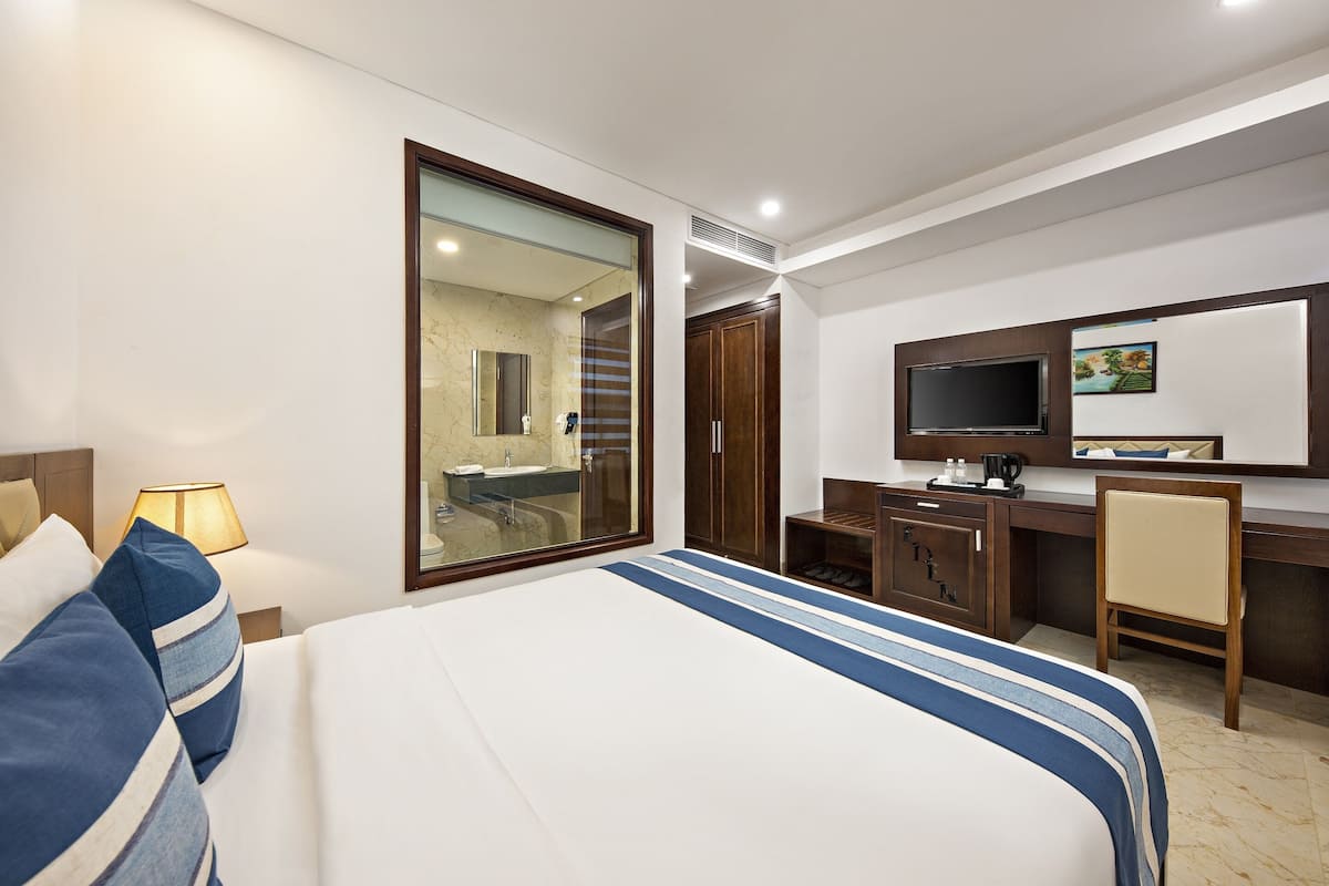 Une chambre d’hôtel avec un grand lit, un bureau avec une chaise, une télévision et une salle de bain visible à travers une porte en verre.