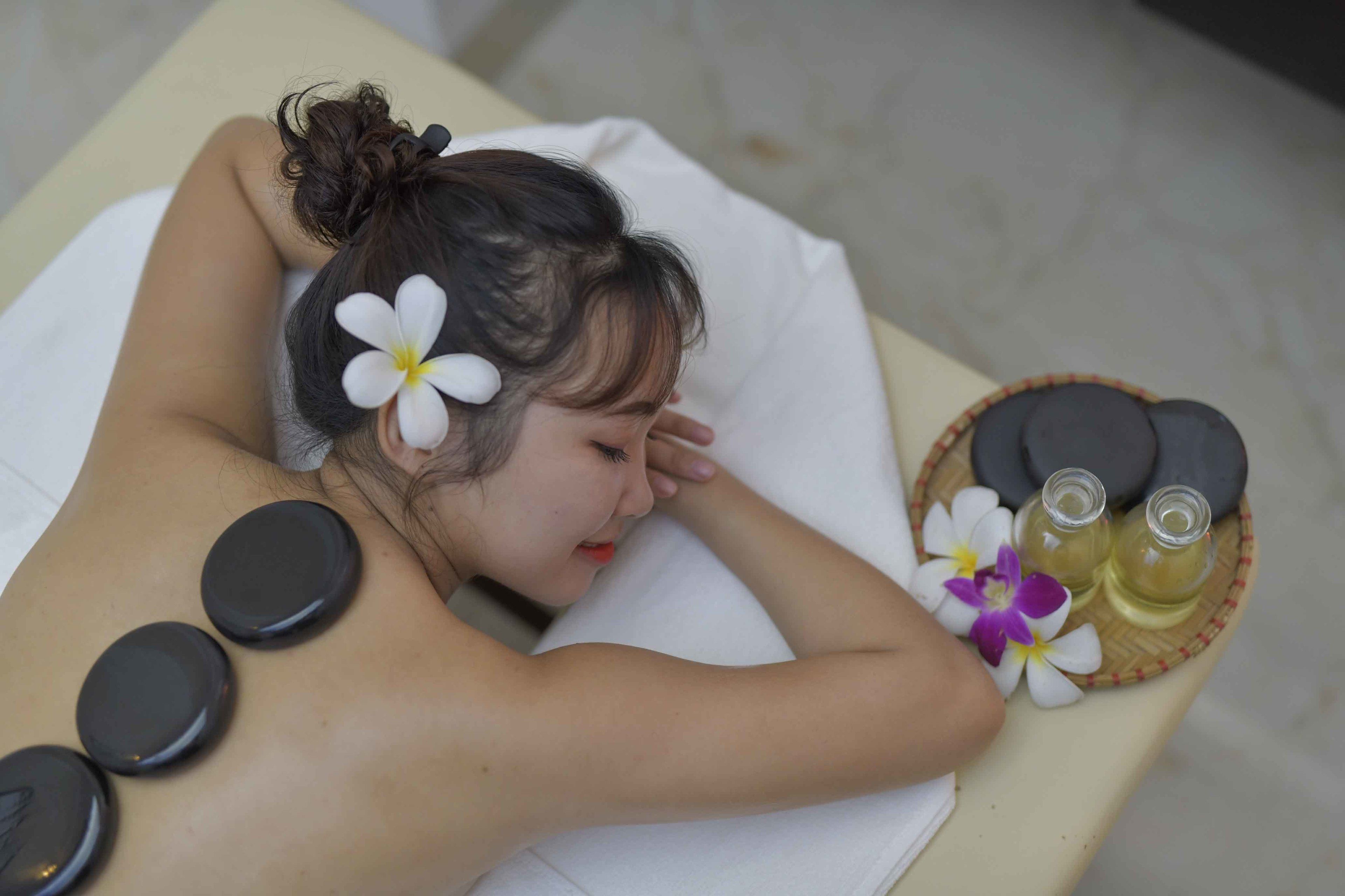 A person receiving a back massage with stones and flowers on a table.