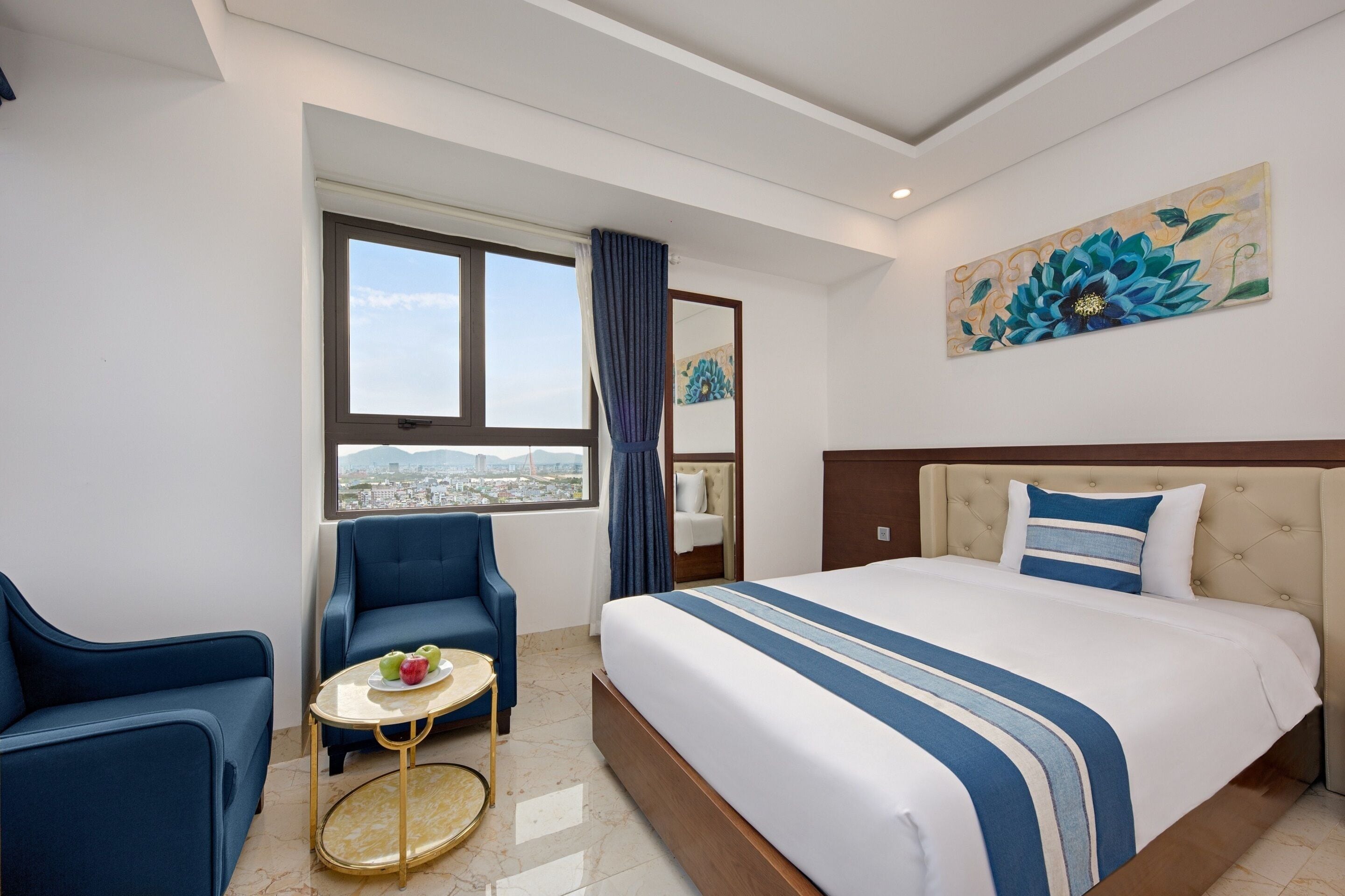 A hotel room with a large bed, a blue armchair, a small round table with fruit, a window with a view of the city, and a floral painting on the wall.