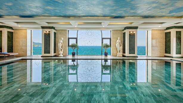 A luxurious indoor swimming pool with a clear view of the ocean through large windows, marble walls, and decorative statues. - Eden Hotel (Da Nang)