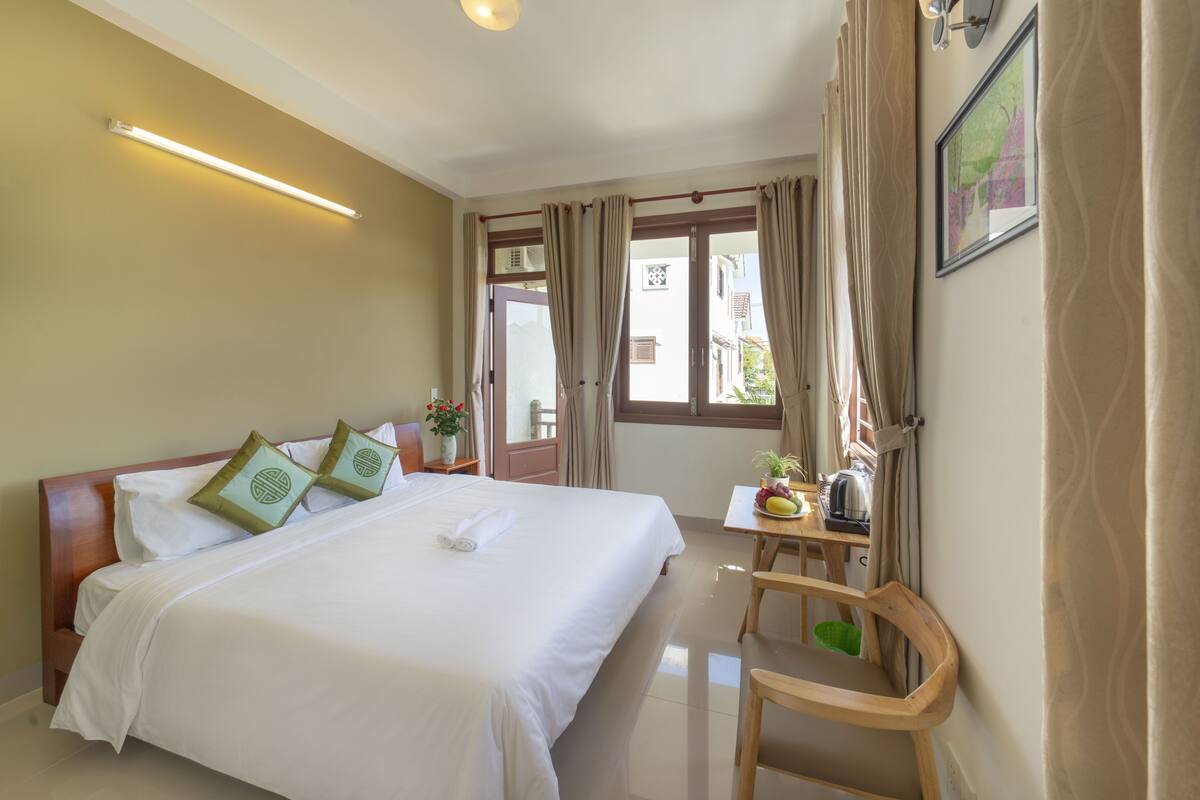 Une chambre d’hôtel avec un grand lit, une petite table sur laquelle se trouve un panier de fruits, une chaise et une fenêtre avec des rideaux.