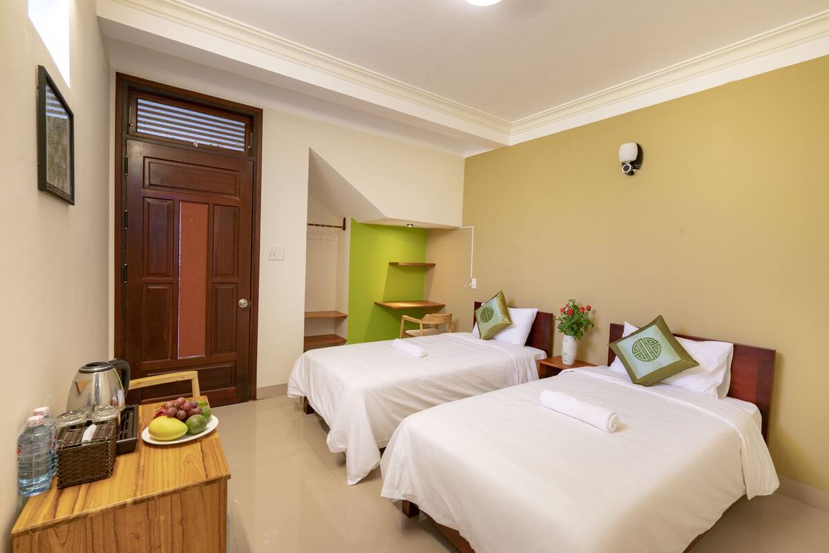 Une chambre d’hôtel avec deux lits, un bureau en bois, une bouteille d’eau, une assiette de fruits et une machine à café.