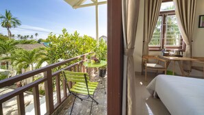 A balcony with a wooden railing, a green chair, a small table with a potted plant, and a view of palm trees and houses. - Green Garden Hoi An Villa (Da Nang)