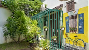 A courtyard with a bench, potted plants, a bicycle, and a building with a yellow wall and blue shutters. - Green Garden Hoi An Villa (Da Nang)