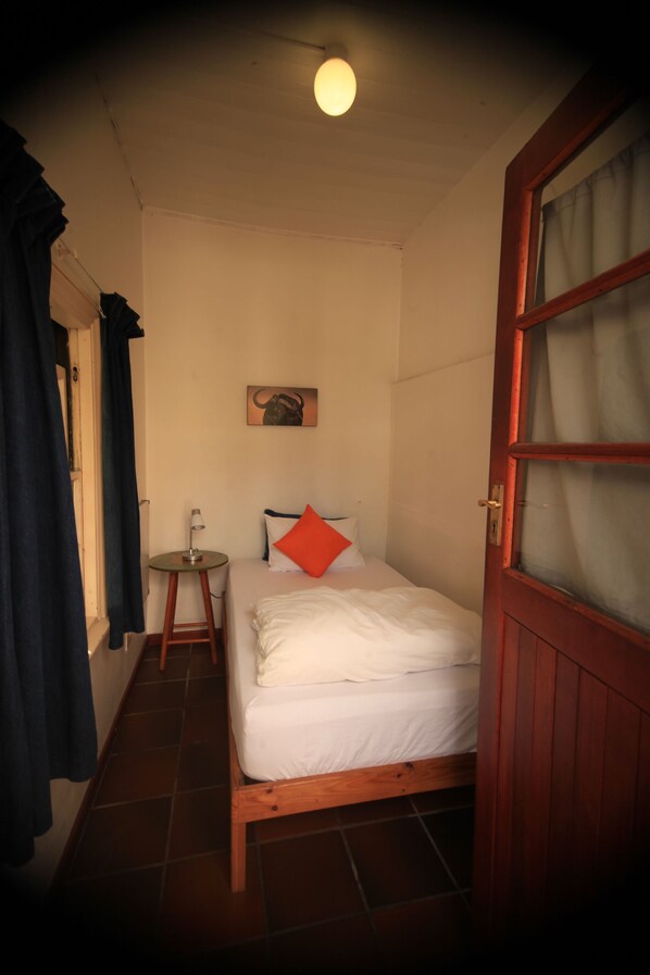 A small, cozy room with a single bed, a small table, and a wall-mounted lamp.