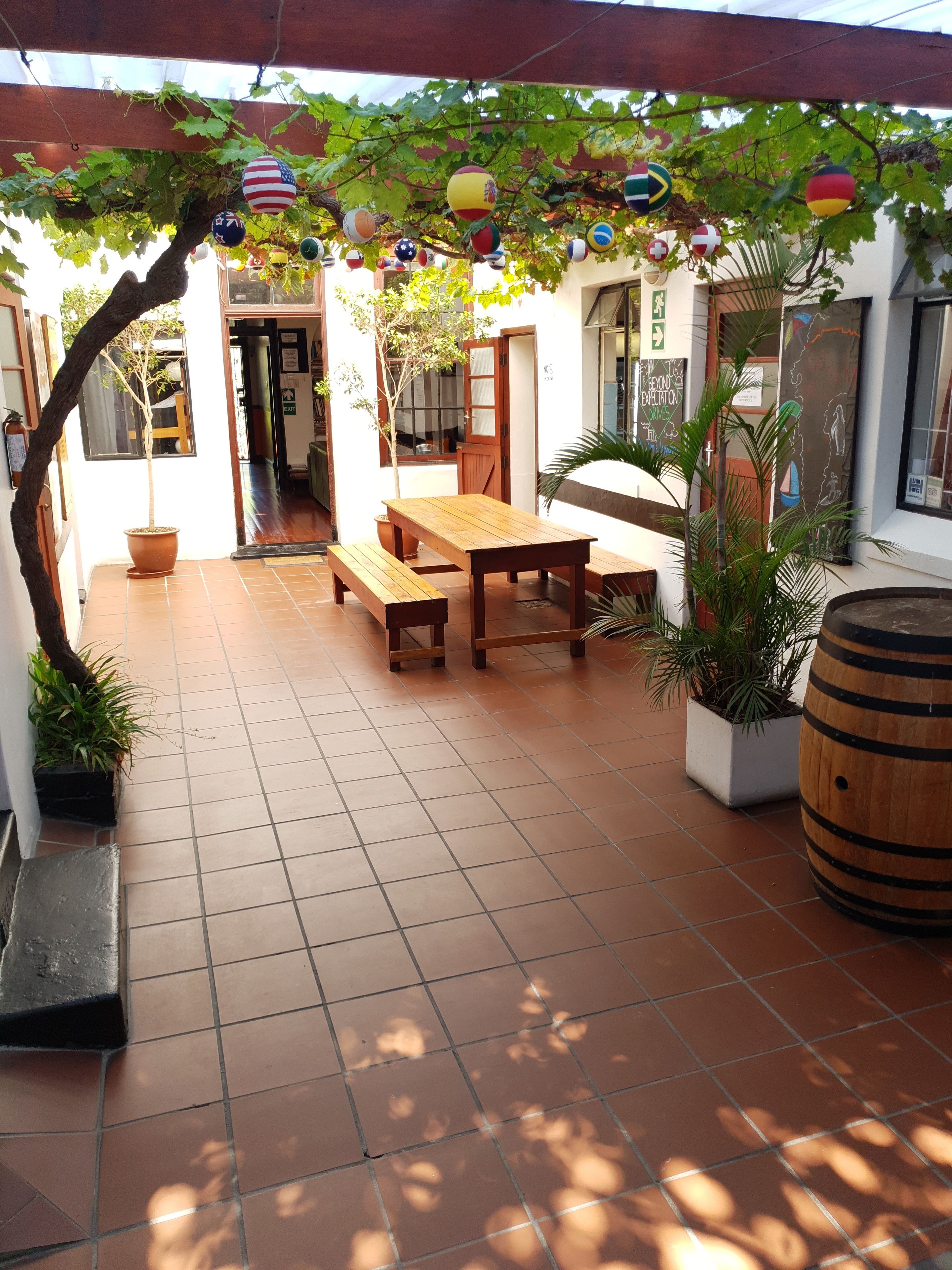 A patio with a wooden table and benches, surrounded by potted plants and a pergola with hanging ornaments.