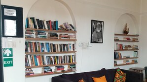 A cozy reading area with a dark-colored sofa, multiple bookshelves filled with books, and a small table with magazines. An exit sign is visible on the wall. - Zebra Crossing Backpackers (Cape Town)