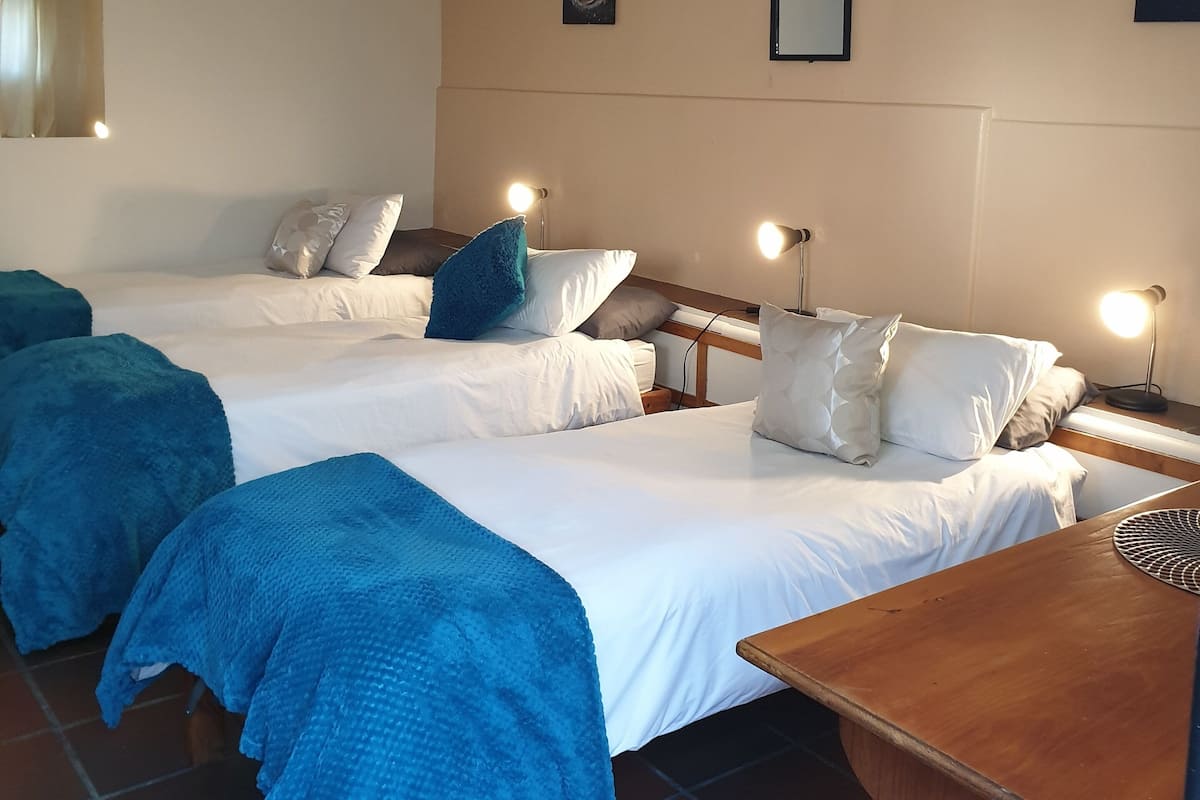 Quarto de hotel com duas camas, cada uma com roupa de cama branca e almofadas azuis. Há mesas de cabeceira com abajures, uma mesinha de madeira e uma pequena mesa de madeira. O quarto tem piso de azulejo e paredes bege.