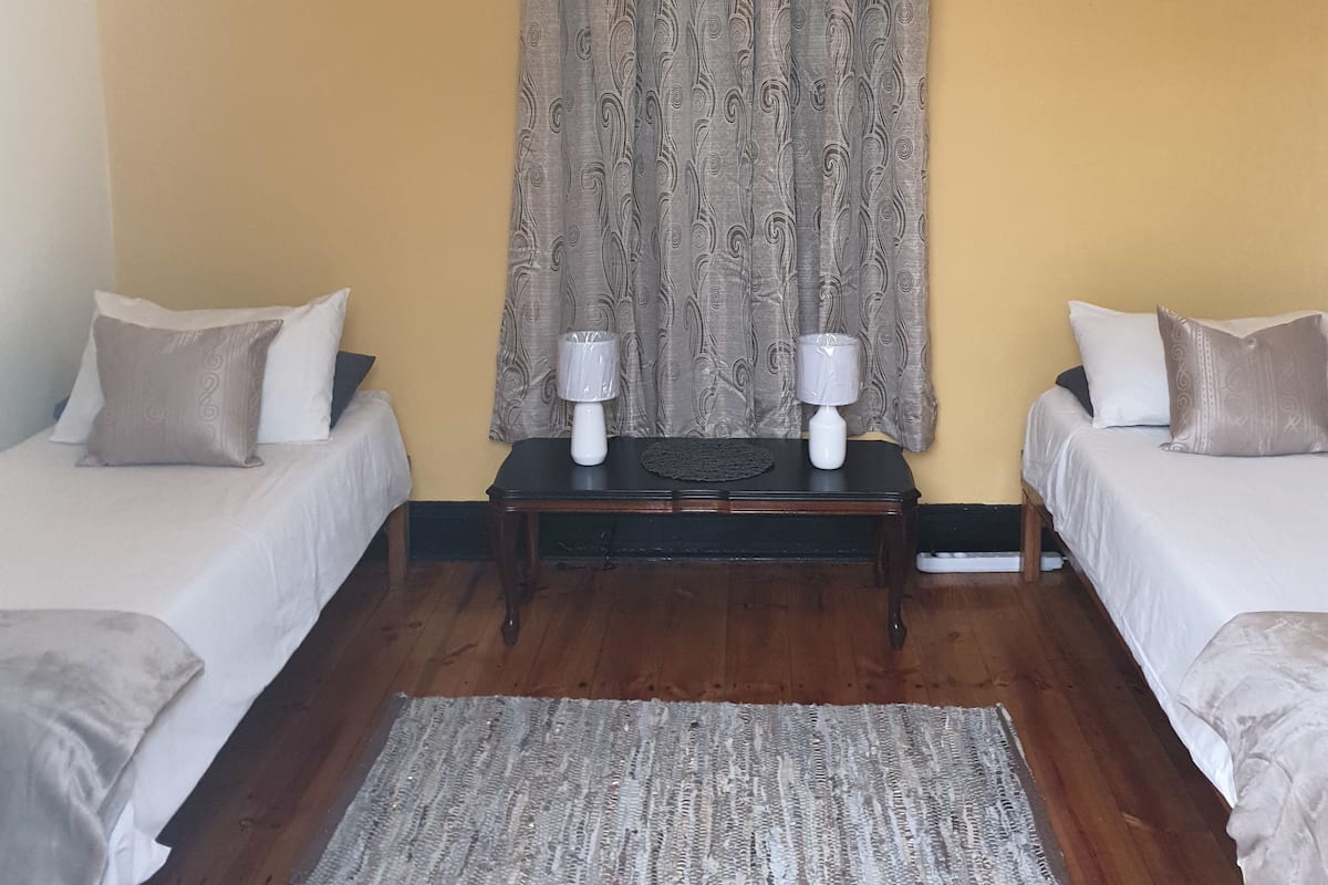 Quarto com duas camas, piso de madeira, cortina estampada e mesa com duas luminárias.