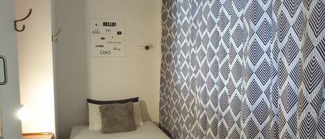 A small, single-bed room with a patterned curtain, a bedside table, and a chair.