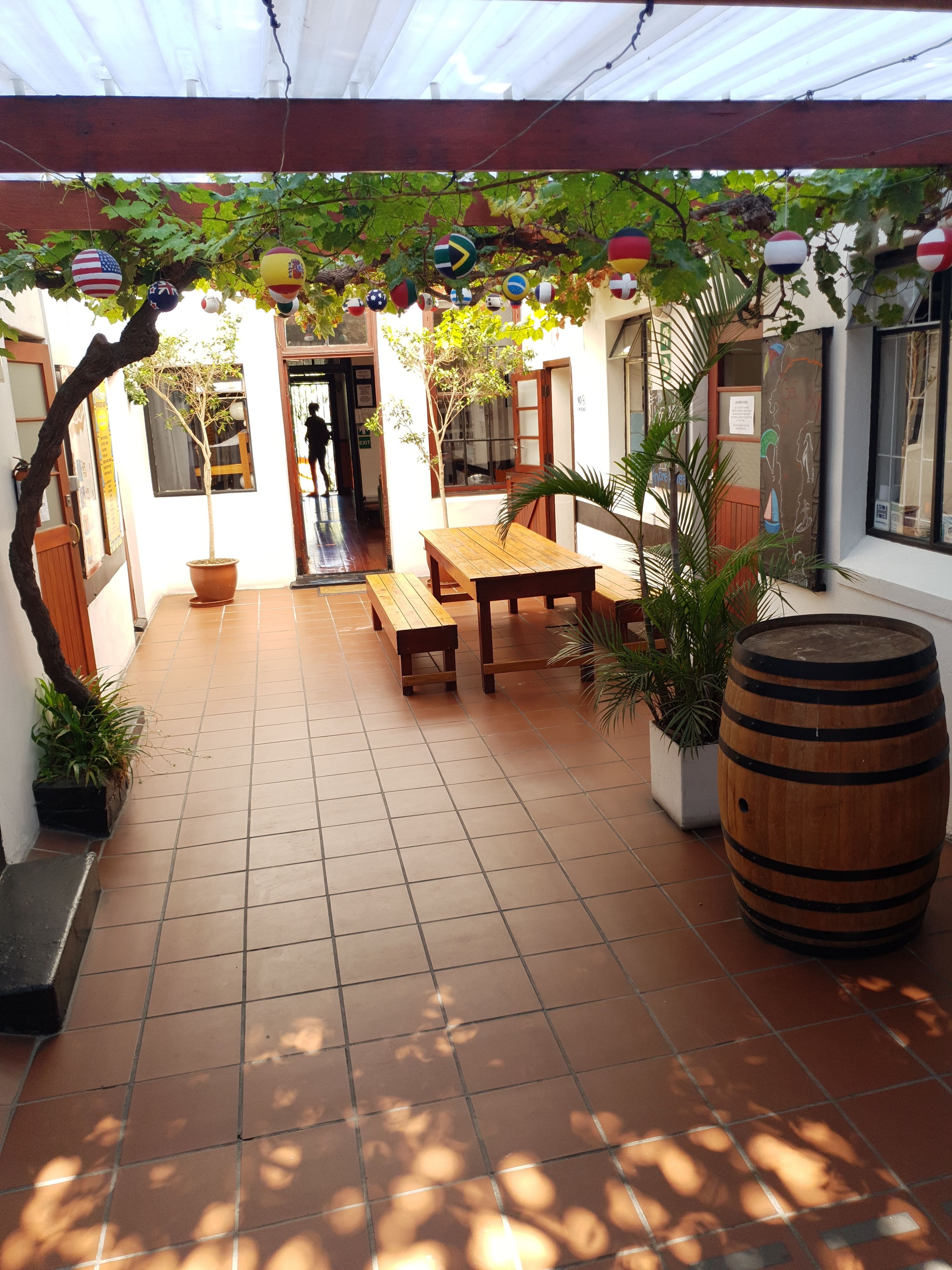 Une terrasse couverte avec une table en bois et des bancs, entourée de plantes en pot et d’un tonneau.