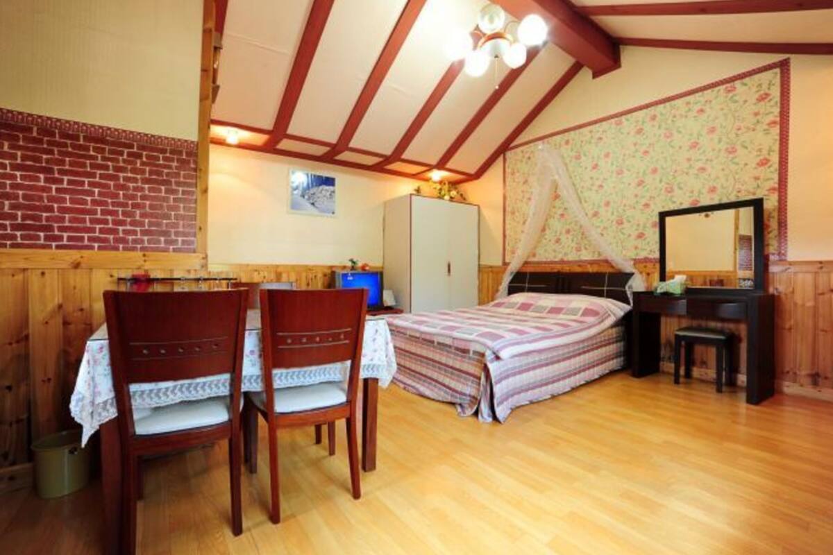 A bedroom with a wooden floor, a bed with a striped bedspread, a desk with a chair, a wardrobe, and a mirror.