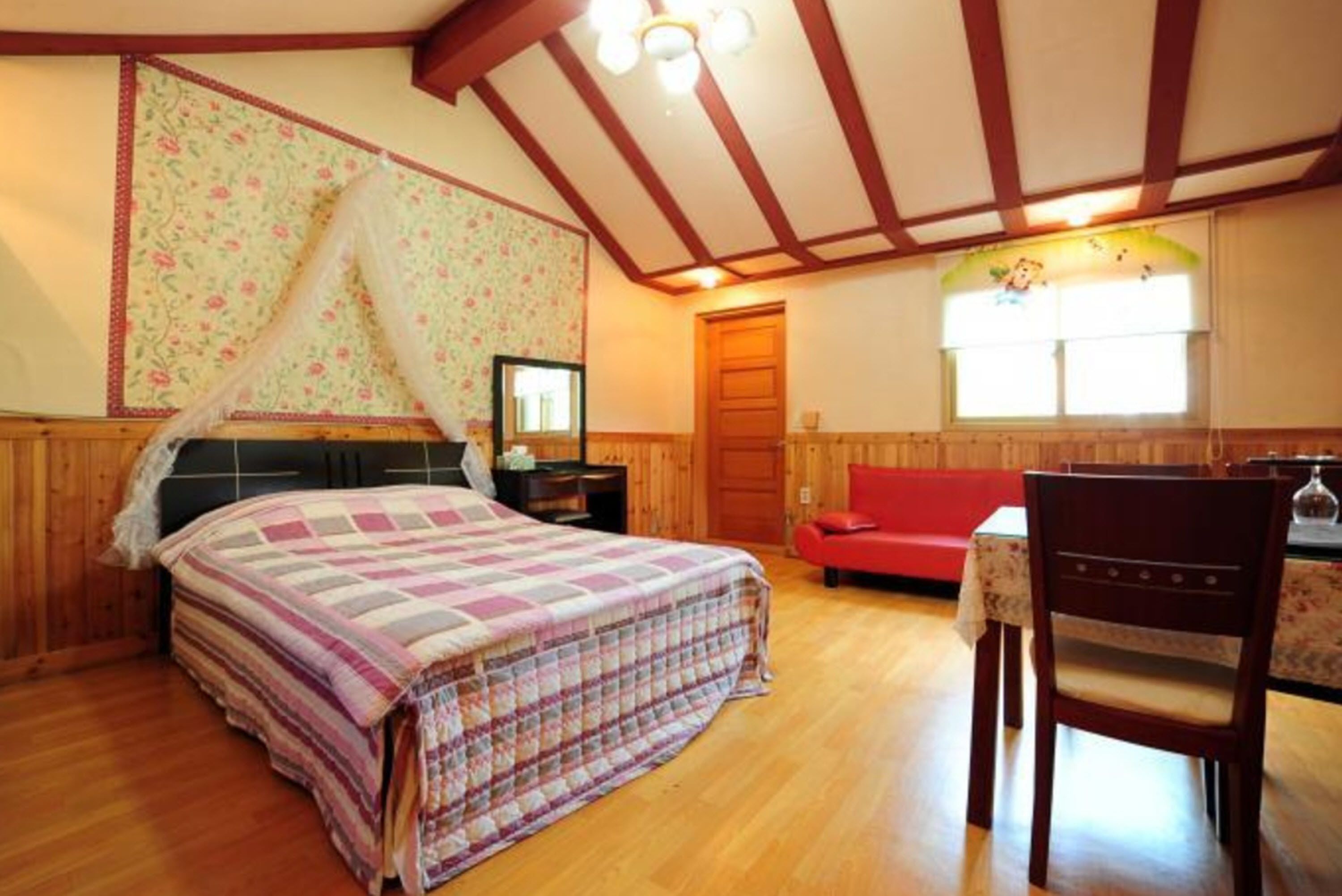 A bedroom with a bed, a nightstand, a red chair, a wooden table, and a window with a floral curtain.