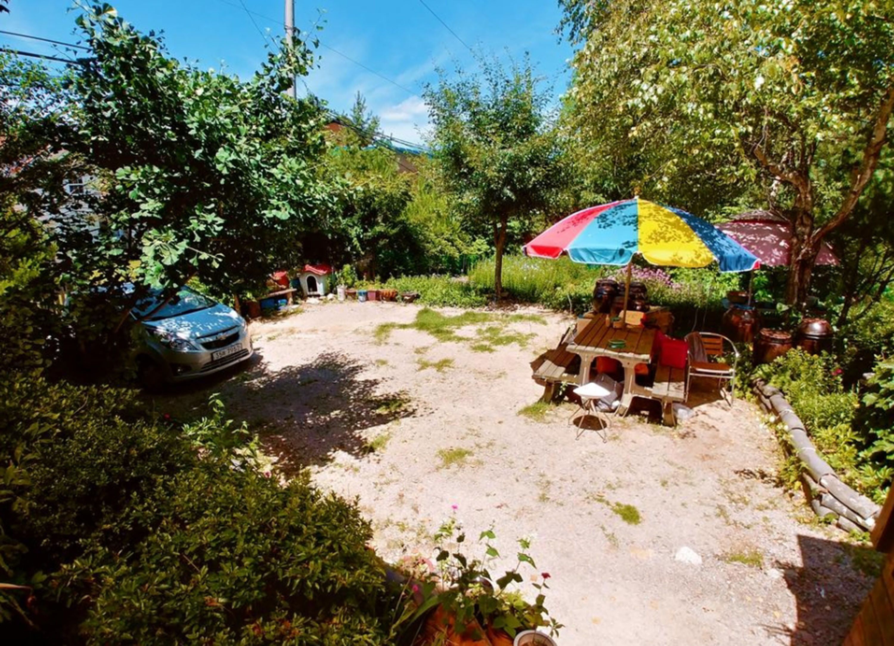 Un espace jardin avec un chemin de terre, une voiture et une table entourée de chaises, le tout sous un parasol coloré.