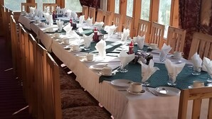 A long dining table set with white napkins, plates, and cups, in a room with wooden chairs and large windows.
